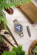 Load image into Gallery viewer, An overhead lifestyle photograph of a Breitling Jupiter Pilot watch centered on a light wood grain surface, framed by green fern leaves and travel accessories.