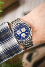 Load image into Gallery viewer, A close-up wrist shot of a person wearing a Breitling Jupiter Pilot chronograph watch with a blue dial, styled with a blue and white checkered flannel shirt.