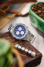 Load image into Gallery viewer, A Breitling Jupiter Pilot watch with a blue dial and steel bracelet resting on a weathered, brown leather-bound book next to a green bowl of roasted coffee beans.