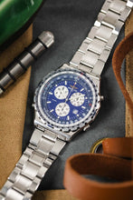 Load image into Gallery viewer, A top-down lifestyle shot of a Breitling Jupiter Pilot watch laying on a grey and tan surface next to a metal spring bar tool and leather straps.