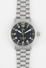 Load image into Gallery viewer, A top-down studio shot of an Omega Dynamic automatic watch with a black dial, white Arabic numerals, and a stainless steel link bracelet. The dial features a yellow center seconds hand and a date window at the 3 o'clock position.