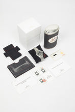 Load image into Gallery viewer, A wide shot of the "full set" including the Omega Dynamic watch on a black cushion, its original white outer box, a unique tin storage case, the international warranty card, and operating instructions manuals.