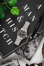 Load image into Gallery viewer, An overhead flat-lay featuring the watch resting on a black book titled "A Man &amp; His Watch," accompanied by a black pen and green leaves.
