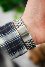 Load image into Gallery viewer, A view of the underside of a wrist showing the brushed stainless steel Omega-branded clasp and the underside of the plaid shirt sleeve.
