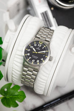 Load image into Gallery viewer, A lifestyle photograph of the Omega Dynamic watch draped over a pair of white leather headphone cushions, with a vintage camera blurred in the background.