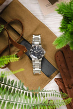 Load image into Gallery viewer, A top-down lifestyle shot featuring an Oris Aquis Date watch on a stainless steel bracelet, centered between a brown leather watch roll, a pair of brown leather gloves, and green foliage.