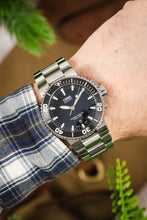 Load image into Gallery viewer, A wrist shot of a person wearing an Oris Aquis Date with a stainless steel bracelet, paired with a blue, grey, and white checkered flannel shirt.
