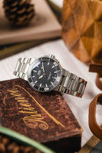 Load image into Gallery viewer, A lifestyle image of an Oris Aquis Date watch with a steel bracelet resting on a vintage brown book titled "Patience," surrounded by leather accents and out-of-focus greenery.