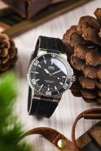 Load image into Gallery viewer, A lifestyle photograph of an Oris Aquis Date watch with a black dial and black rubber strap, positioned next to a large brown pinecone and a leather strap on a light wood surface.